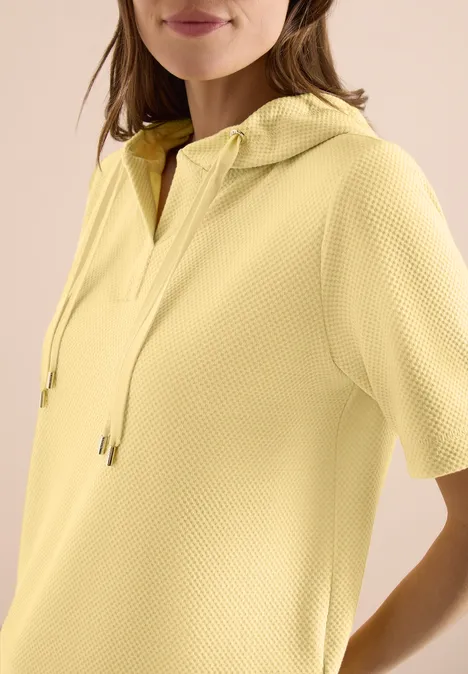 Frau in gelbem, kurzärmeligen Kapuzenpullover mit feiner Struktur.