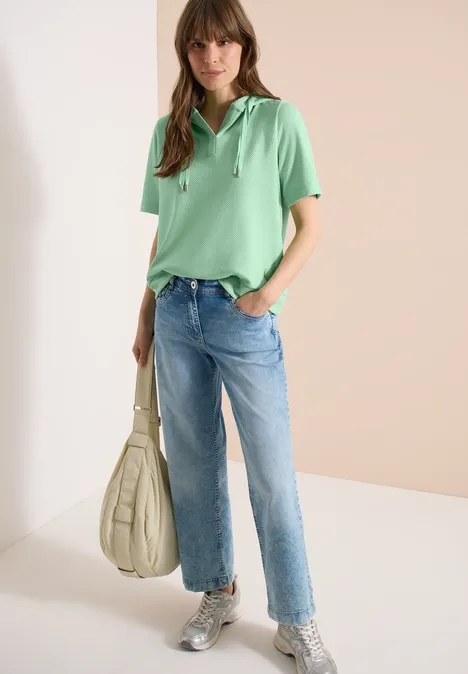 Frau trägt grünes Poloshirt, gerade Jeans und graue Sneaker, hält weiße Beuteltasche vor hellem Hintergrund.