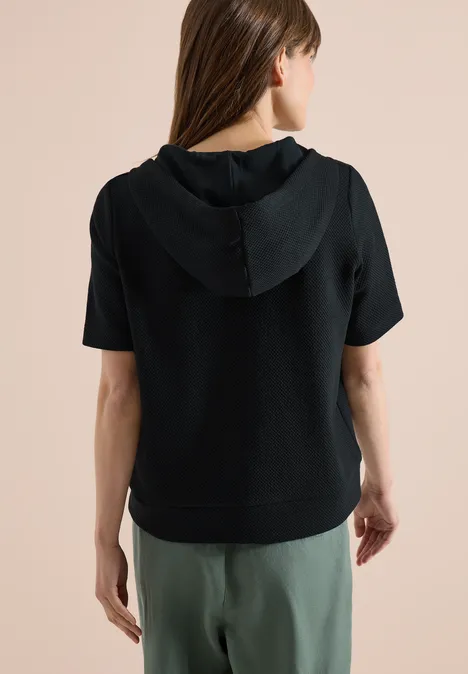 Frau in schwarzem kurzärmligem Hoodie und grüner Hose, Rückenansicht vor hellem Hintergrund.