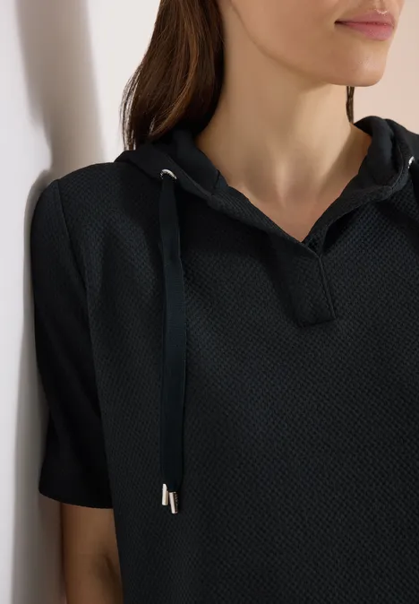 Frau im Profil, lehnt an einer Wand und trägt ein schwarzes kurzärmeliges Kapuzen-Shirt.