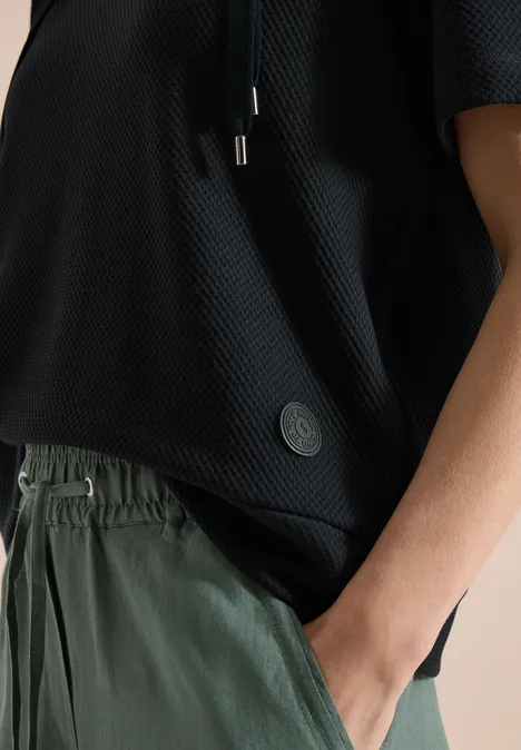 Detailaufnahme einer Person in schwarzem, leicht transparentem Oberteil und grüner Hose mit Hand in der Tasche.