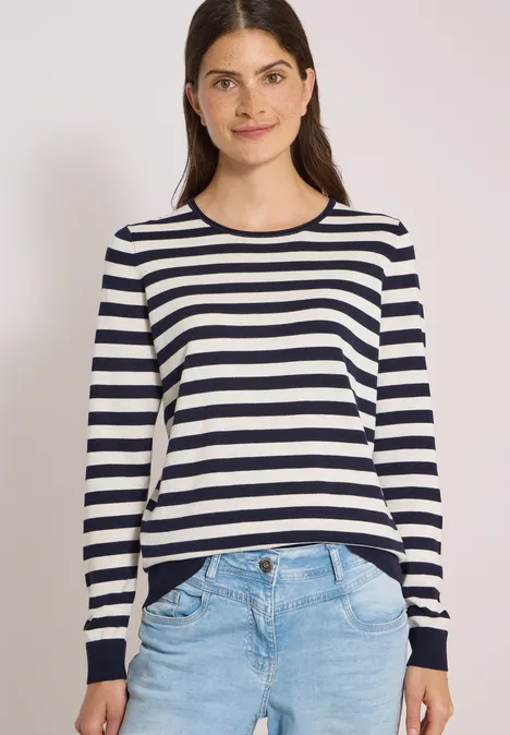 Pull à col rond et rayé urban dark blue Junge Frau in gestreiftem Langarmshirt und Jeans, lächelt vor weißem Hintergrund.