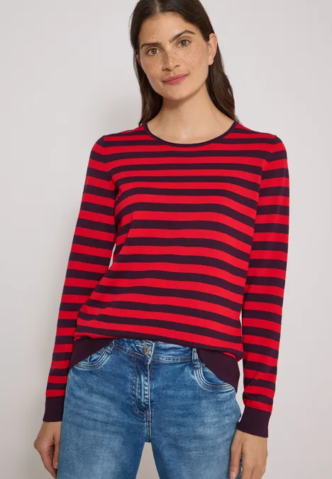 Pull à col rond et rayé eggplant red Frau trägt rotschwarz gestreiftes Langarmshirt und blaue Jeans.