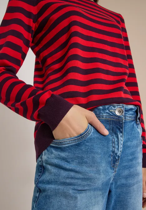 Pull à col rond et rayé eggplant red Roter, schwarz gestreifter Pullover mit langen Ärmeln und blaue Jeans, eine Hand in der Hosentasche.