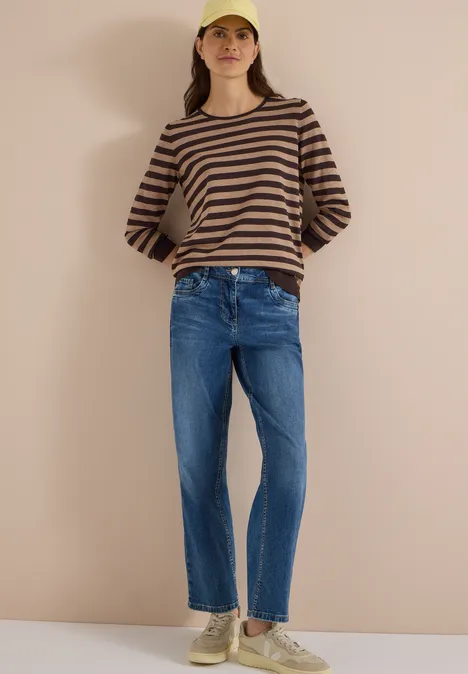 Pull à col rond et rayé truffle brown Frau in gestreiftem Pullover, Jeans, Sneakern und gelber Mütze vor neutralem Hintergrund.