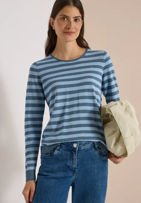 Jeune femme en T-shirt bleu à rayures, jean, tenant une veste matelassée beige clair sous le bras.