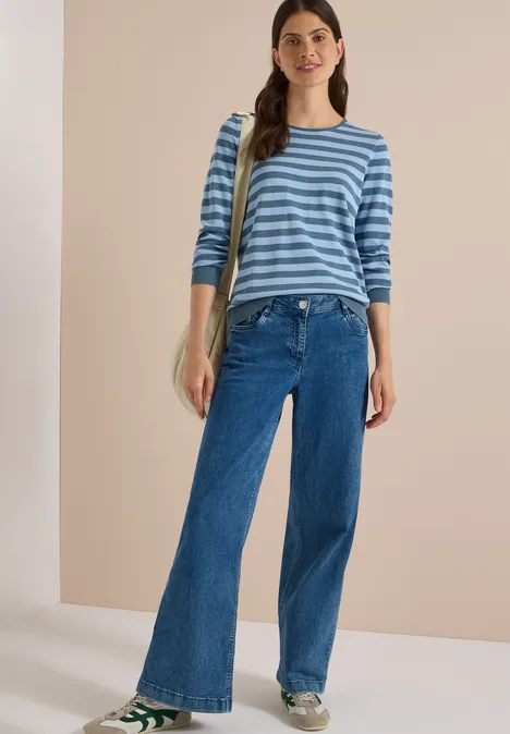 Une femme porte un pull rayé bleu et blanc, jean large et baskets, posture décontractée avec sac.