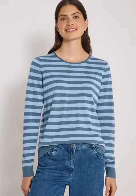 Femme aux cheveux brun foncé porte un t-shirt à manches longues bleu à rayures et un jean, souriante sur fond blanc.