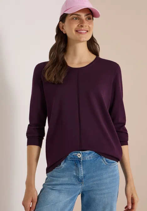 Lächelnde Frau mit lila Pullover, Jeans und rosa Cap vor neutralem Hintergrund.