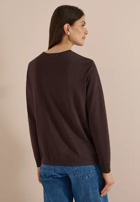 Eine Frau im Profil trägt einen schlichten dunkelbraunen Pullover und blaue Jeans vor hellem Hintergrund.