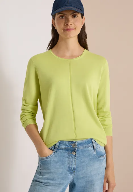 Frau trägt limettengrünen Pullover und blaue Jeans, Hände in den Taschen, mit dunkler Baseballmütze.