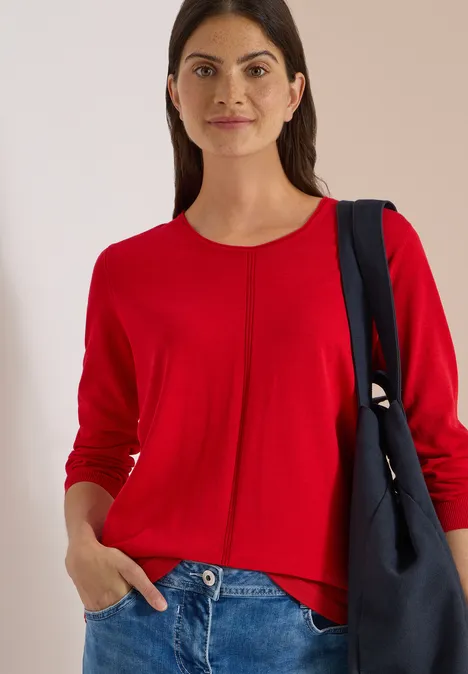 Frau in rotem Pullover und Jeans, lächelnd, Hand in Tasche, mit Schultertasche.