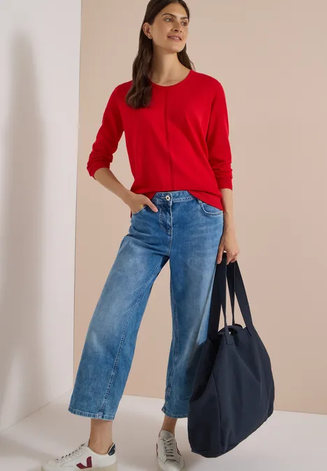 Frau in rotem Pullover, weiten Jeans und weißen Sneakern, die eine große blaue Tasche trägt.