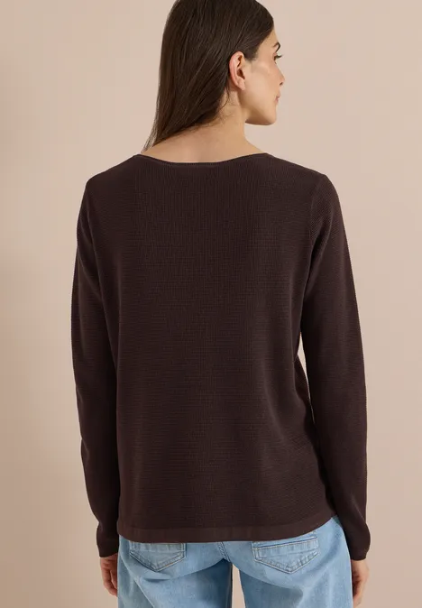 Rückansicht einer Frau in einem dunkelbraunen Langarmshirt und blauen Jeans vor hellem Hintergrund.
