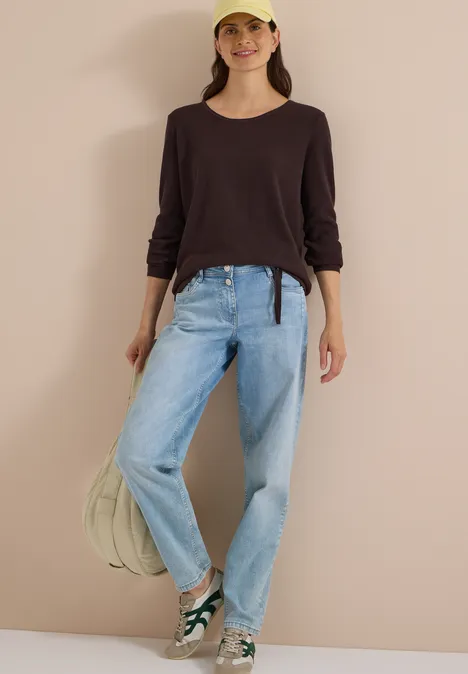 Junge Frau in lässigem Outfit mit Basecap, dunklem Oberteil, hellen Jeans und Sneakers, stehend vor hellem Hintergrund.