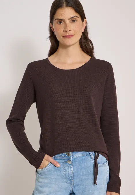 Frau mit dunklen Haaren in braunem Langarmpullover mit Bindeband und Jeans.