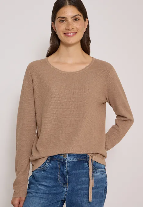Junge Frau mit langem dunkelbraunem Haar trägt einen beigen Pullover und blaue Jeans.