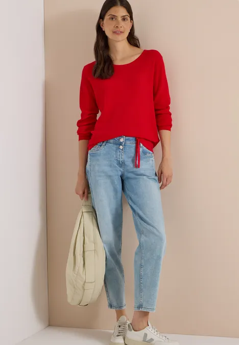 Eine Frau trägt roten Pullover, helle Jeans, graue Sneakers und hält eine beigefarbene Tasche.