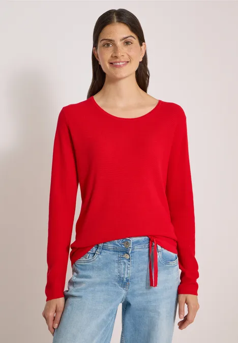 Junge Frau lächelt, trägt roten Pullover mit Knoten am Saum und hellblaue Jeans vor einfachem Hintergrund.