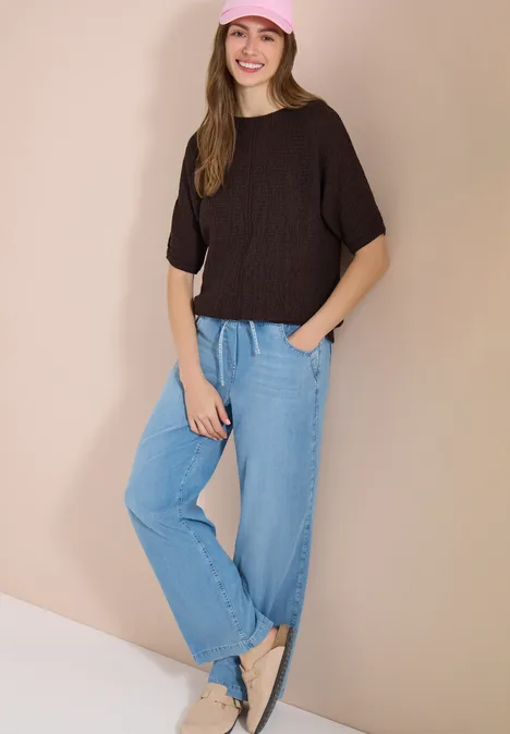 Dolman Pullover mit U-Boot-Ausschnitt brown cacao Eine Frau lehnt an einer beigen Wand, trägt schwarzen Pullover, lockere hellblaue Jeans und beige flache Schuhe.