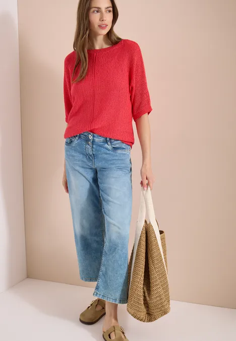 Femme en haut rouge à manches 3/4, jean large court et mocassins, avec un grand sac en paille.