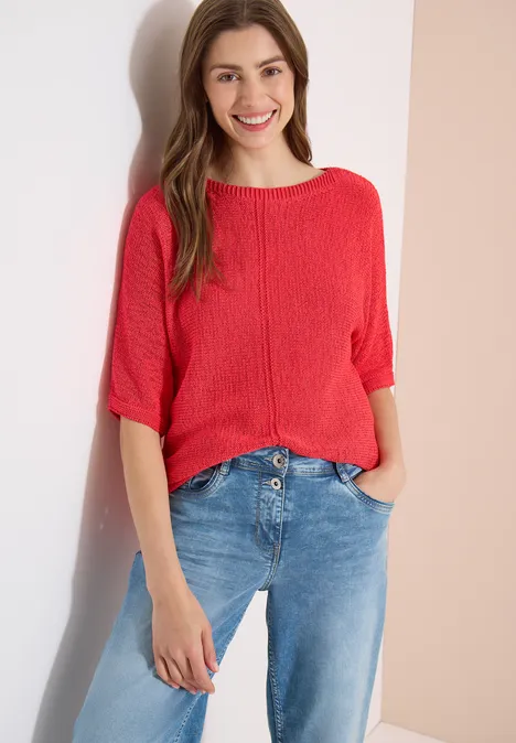Femme appuyée contre un mur, porte un haut rouge ample et un jean bleu.