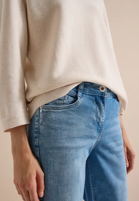 Person in beigem Oberteil und blauen Jeans, Fokus auf Oberkörper mit entspannter Hand.