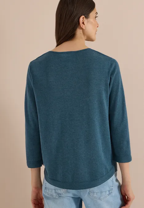 Frau von hinten in blau-grünem Langarmshirt und blauer Jeans vor neutralem Hintergrund
