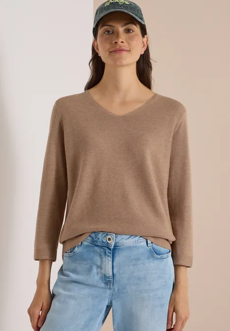Frau trägt blaue Jeans, beigefarbenen V-Pullover und Cap, steht vor hellem Hintergrund.