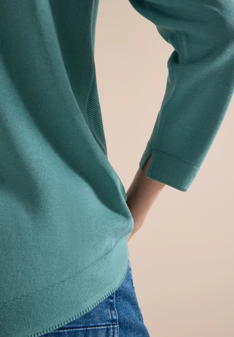 Nahaufnahme eines türkisfarbenen Langarmshirts mit seitlichem Schlitz am Ärmel über blauer Jeans.