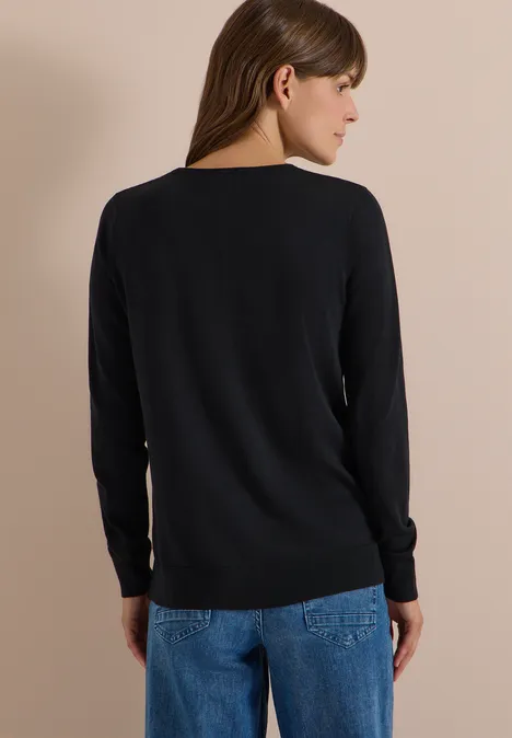 Eine Frau steht mit dem Rücken zur Kamera, trägt schwarzen Pullover und blaue Jeans vor hellbeigem Hintergrund.