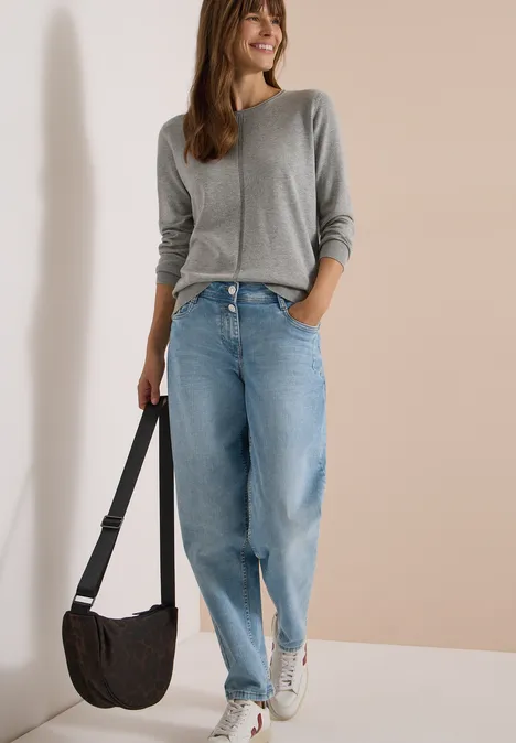Jeune femme en pull gris, jeans larges et baskets blanches, avec un sac bandoulière, sur fond neutre.