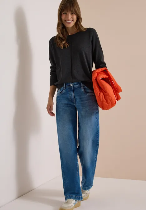 Femme souriante en tee-shirt noir à manches longues et jean bleu tenant une doudoune orange sous le bras.