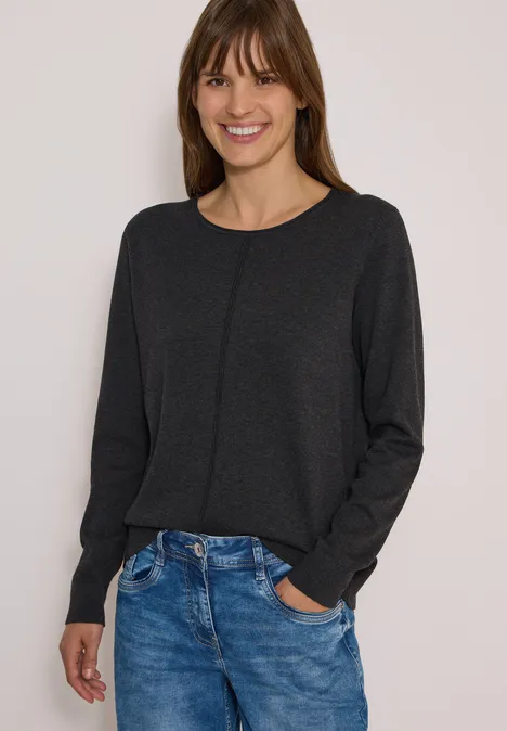 Femme souriante en pull gris foncé et jean bleu, posant devant un fond clair.
