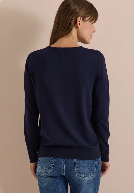 Eine Frau von hinten in dunkelblauem Pullover und Jeans vor beigem Hintergrund.