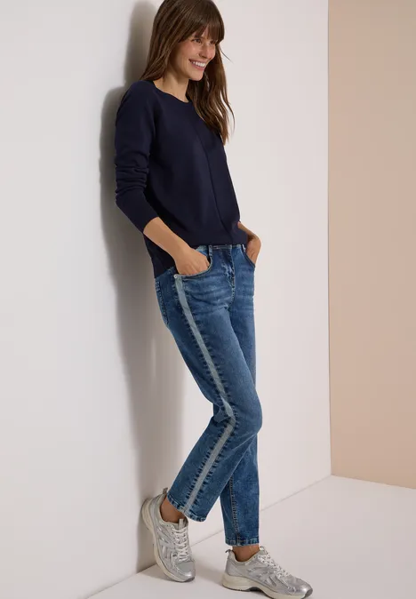 Frau lehnt lässig in Jeans, dunkelblauem Pullover und Sneakern an einer Wand und lächelt.