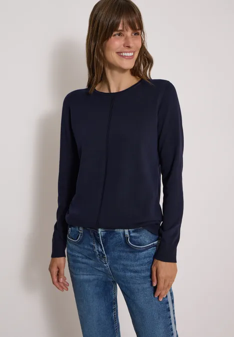 Frau in dunkelblauem Pullover und Jeans steht lächelnd vor hellem Hintergrund.