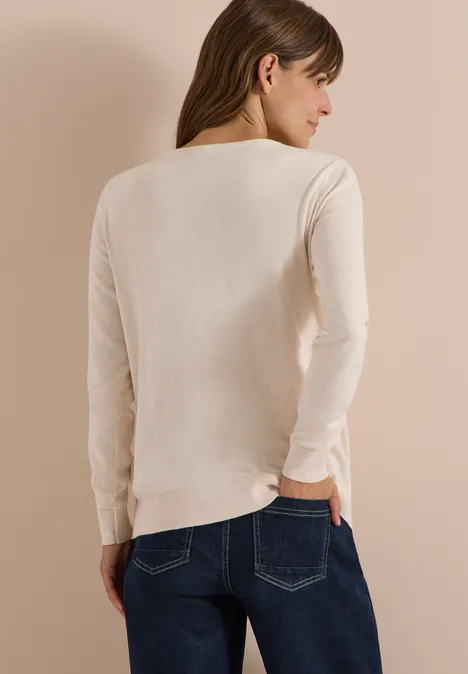 Eine Frau von hinten in einem hellbeigen Langarmpullover und blauer Jeans vor hellem Hintergrund.