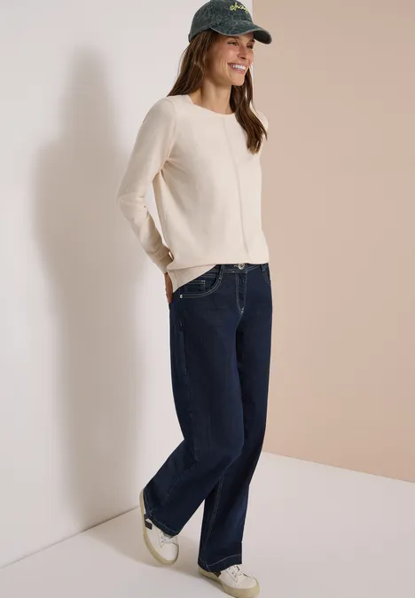 Frau in beigem Pullover, breiten Jeans, weißen Sneakern und grüner Baseballkappe lehnt lächelnd in der Ecke.