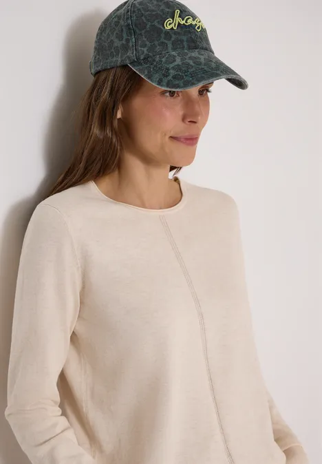 Eine junge Frau trägt einen hellbeigen Pullover mit mittiger Naht und eine dunkelgrüne Cap mit Muster.