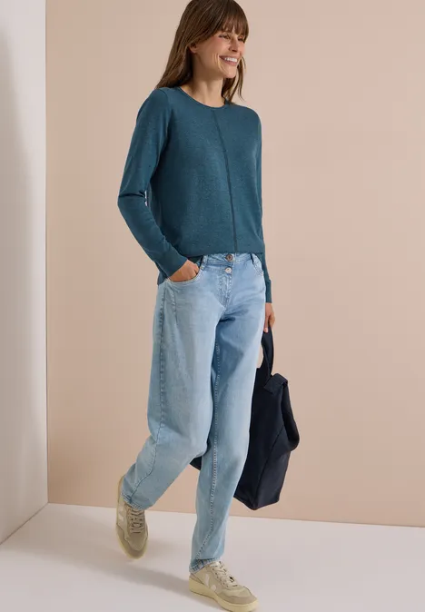 Eine Frau in locker sitzender Jeans, tealfarbenem Pullover und hellen Sneakern, die lächelnd eine Jacke hält.