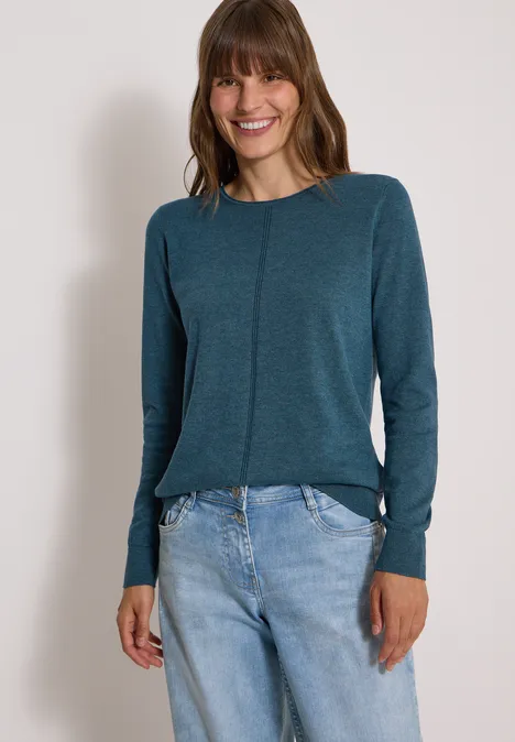 Eine lächelnde Frau in einem petrolfarbenen Langarmshirt und hellen Jeans vor neutralem Hintergrund.