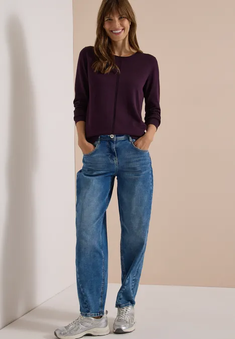 Femme détendue appuyée contre un mur, pull violet, jean bleu ample et baskets argentées.