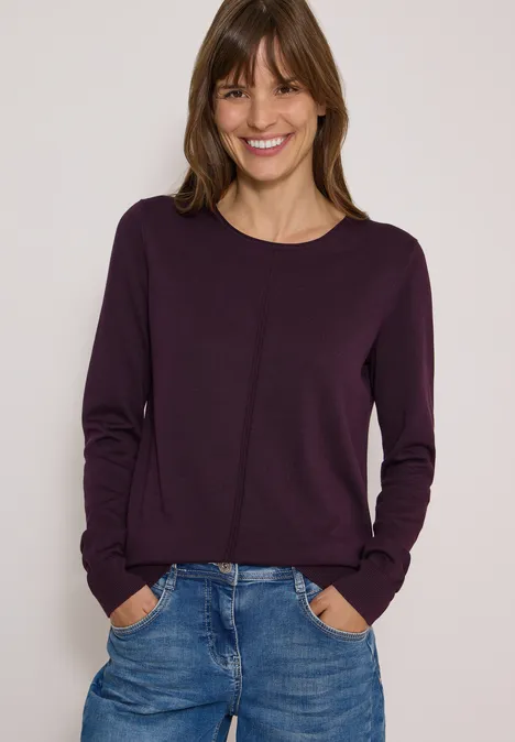 Femme souriante en pull violet et jeans, mains dans les poches, sur fond clair.