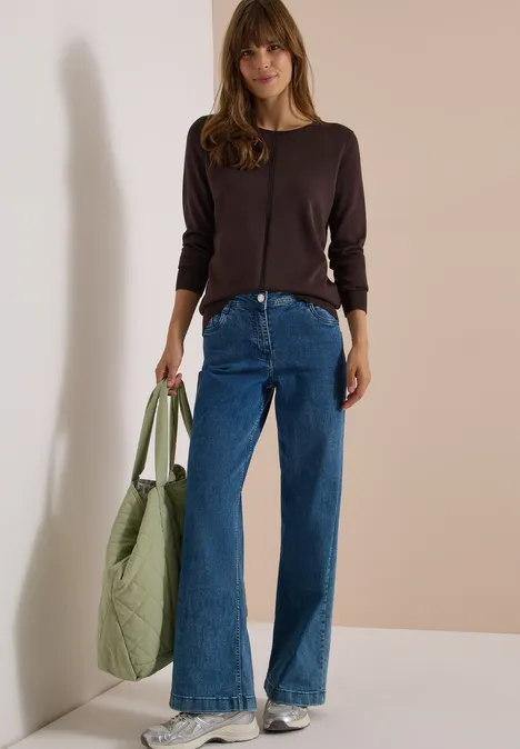Une femme posant contre un mur, en jean bleu large, haut marron, baskets et grand sac vert matelassé.