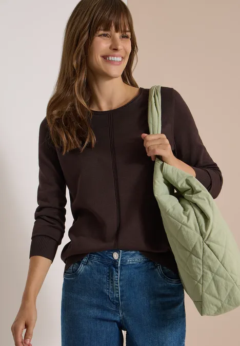 Femme en pull marron et jean, souriante, portant un sac matelassé vert clair à l'épaule.