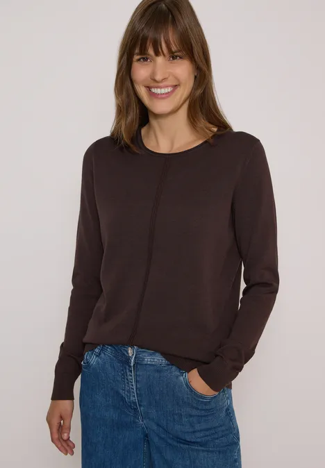 Femme souriante en pull marron et jean devant un fond clair.