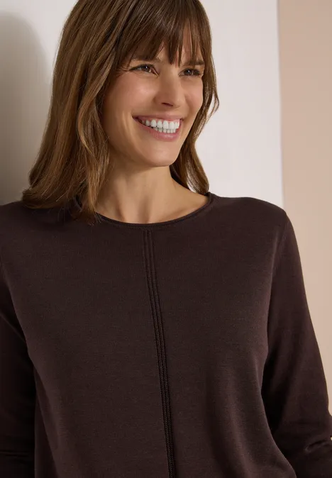Femme souriante en pull marron devant un mur clair.
