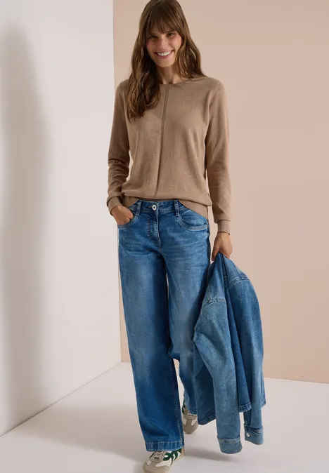 Frau im beigen Cardigan und weiten Jeans, hält eine Jeansjacke in der Hand vor hellem Hintergrund.