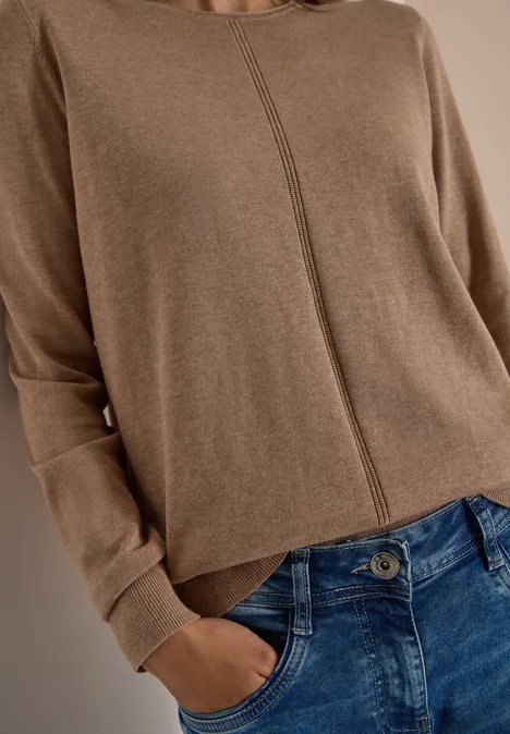Person trägt braunen Pullover mit Mittelnaht und blaue Jeans, eine Hand in der Hosentasche.